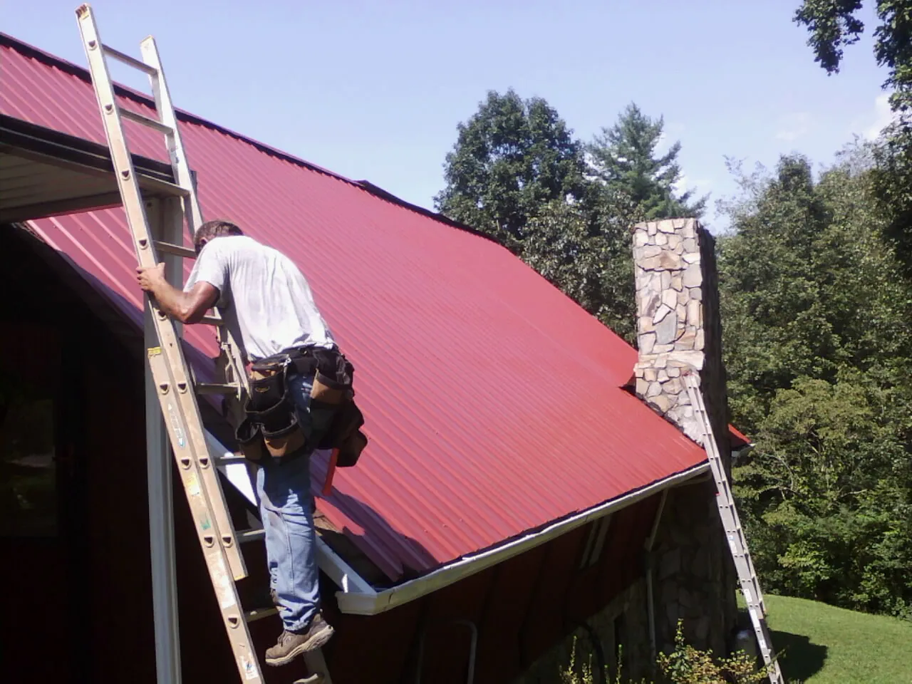 Roofer on a ladder beside a red metal roof and stone chimney — Soffit & Fascia Repair by Roofing Brothers in Blue Ridge Summit